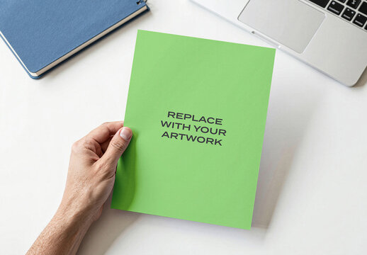Square brochure mockup held in hand on white desk with laptop and notebook, realistic print presentation for graphic design, branding, advertising, editorial, stationery marketing materials