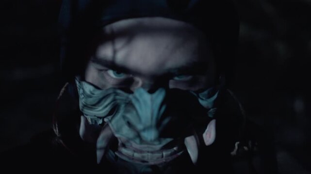 Tight frontal close-up shows person wearing weathered painted mask with exaggerated nose and carved teeth, eyes visible above mask, subtle head tilt, tense watchful expression, low-key outdoor light