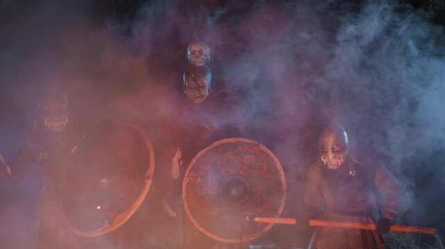 Three costumed percussionists perform facing forward on stage while striking large round gongs with wooden mallets, surrounded by colored smoke, bright reflections, and intense lighting that creates