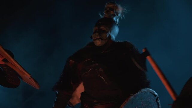 Group of masked performers hold carved round shields and drumsticks while leaning forward in poised rhythm, with raised arms and smoky blue backlight accented by red rim glow for dramatic impact