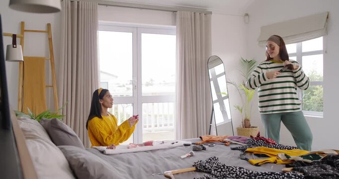 Diverse women sorting clothes in bedroom, holding polka-dot dress, mirror near, planning outfits