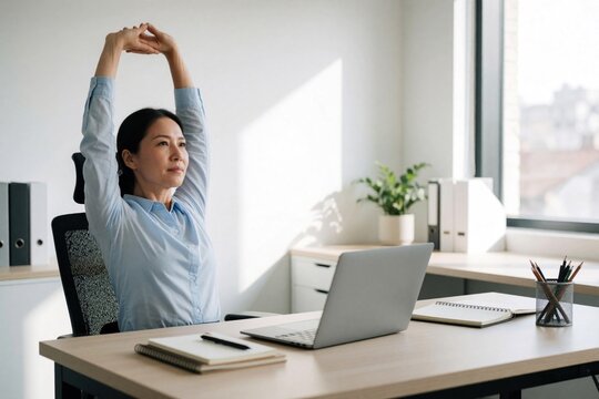 Asian woman stretches arms up while sitting at desk in office. Employee combats fatigue with movement, improves posture and relieves tension from prolonged work.