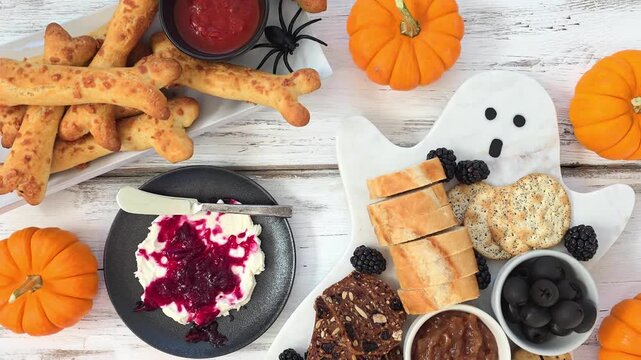 Spooky Halloween grazing table scene with bone breads, ghost charcuterie board and blood splatter cream cheese. Overhead view on a white wood background. Slow handheld panning motion.