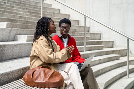African american friends man and woman students talking sharing ideas while working outside on laptop after college classes, confident intelligent black male and female brainstorming and interacting.