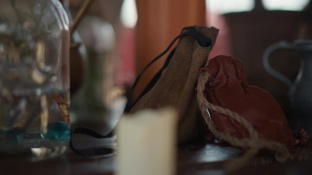 Close view of worn leather pouch with frayed cord, slightly tilted against dried red leaf on wooden tabletop, with warm candle glow, blurred glass bottle, soft shadow, and rustic natural textures