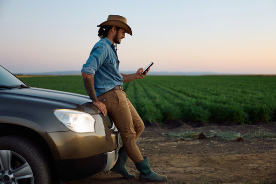 Happy farmer leaning on his truck while texting on smart phone in field.