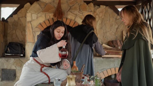 Seated woman raises goblet toward standing companion who pours red drink from small flask, while third woman tends oven nearby in warm stone kitchen with rustic table, pottery, and firelight