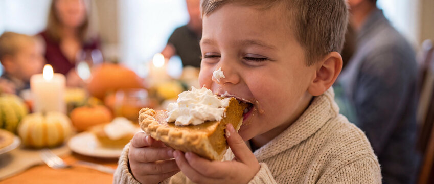 Happy young boy eating a large slice of pumpkin pie with whipped cream during a festive family Thanksgiving dinner
