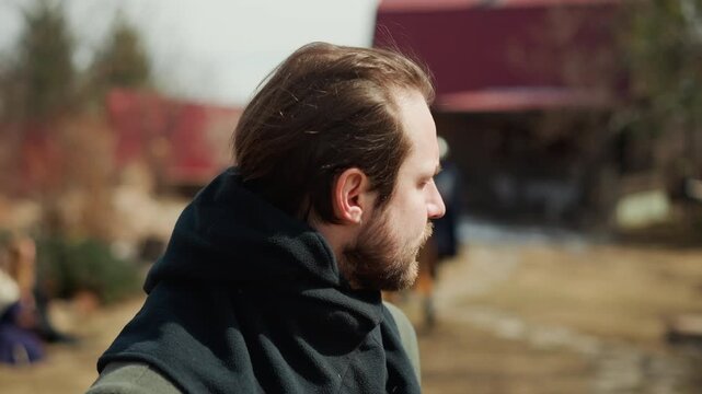 Three-quarter side portrait of adult bearded man walking outdoors with focused gaze, slight lean, and calm expression, wearing casual coat and scarf beside softly blurred urban street scenery in