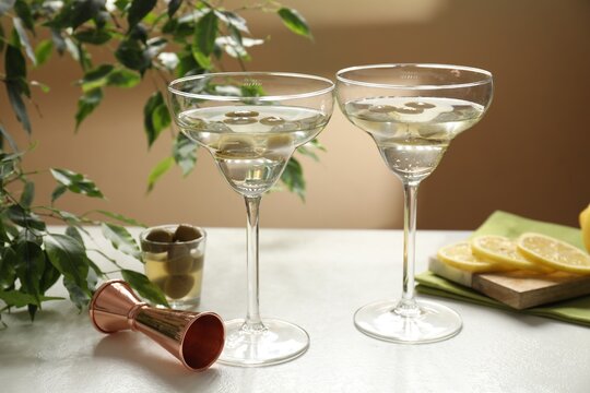 Martini cocktails with olives in glasses, slices of lemon, jigger and green plant on white table, closeup