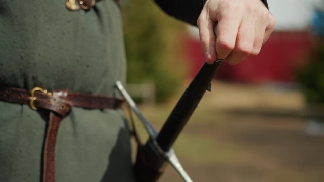 Waist-level close-up shows hands drawing sword by hilt and pommel, fingers firm on grip and wrist turned slightly, while wool tunic and leather belt suggest historic outdoor reenactment