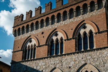 Fototapeta premium Brick, stone medieval building facade with arched windows, crenellations. Historic architecture detail in old European town under blue sky with clouds. Exterior view of ancient Italian structure.