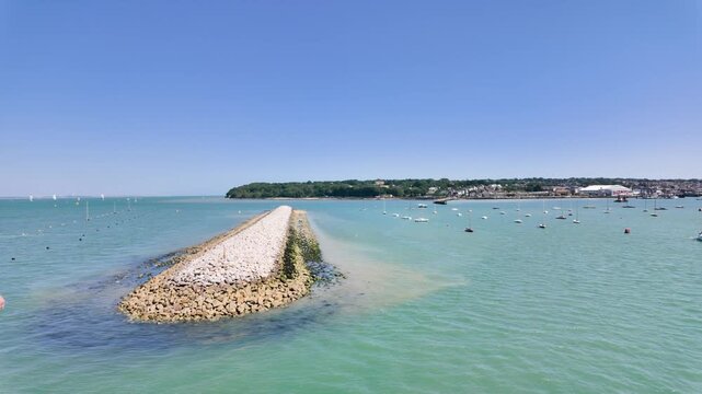 Slow motion footage from an Isle of Wight ferry showing a long stone breakwater stretching through turquoise coastal water with moored sailing boats and the island shoreline beneath a clear blue sky