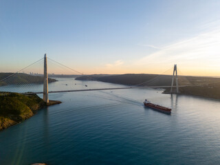 Fototapeta premium Aerial vertical view of Yavuz Sultan Selim Bridge spanning Bosphorus Strait in Istanbul, Turkey. Modern cable-stayed bridge with cargo ship navigating through sea at golden sunset.