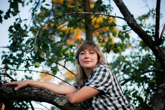 Portrait of a woman leaning on a tree branch in an apple orchard and looking at the camera