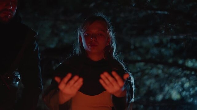 Woman stands outdoors at night with both hands extended forward, palms outward and fingers spread in stopping or shielding gesture; wet hair, damp clothing, warm orange light, and dark forest