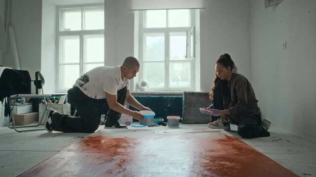 Caucasian young adult male and female artists collaborating on large canvas on floor, pouring paint in studio. Clip is perfect for creative agency promos and art school advertising