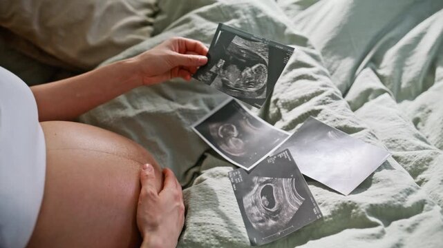 Expectant mother examining ultrasound scans on bed. Closeup hand touching belly.