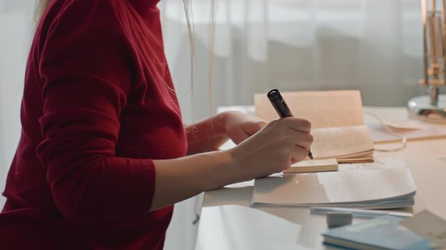 Young woman in red top writes on sticky notes and organizes papers. Woman uses black marker to mark cards labeled. Scene is well lit by natural light through sheer curtains