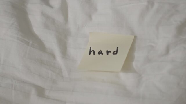 Close up of yellow sticky note with handwritten word hard. Camera focuses steadily as light and shadow subtly shift revealing. Slow zoom in gradually brings details of both text