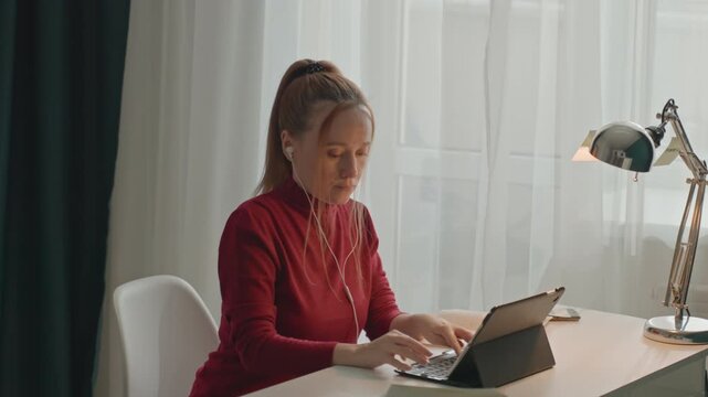Young woman in red long sleeve top types on tablet at white desk. Silver adjustable lamp sits. Camera remains mostly stationary capturing consistent activity showing woman