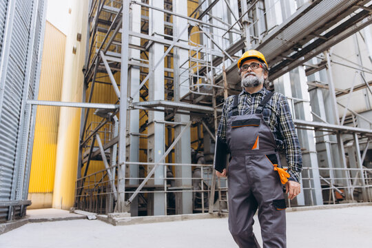 Agro granary elevator worker inspecting industrial agricultural plant with grain silos and complex steel structures