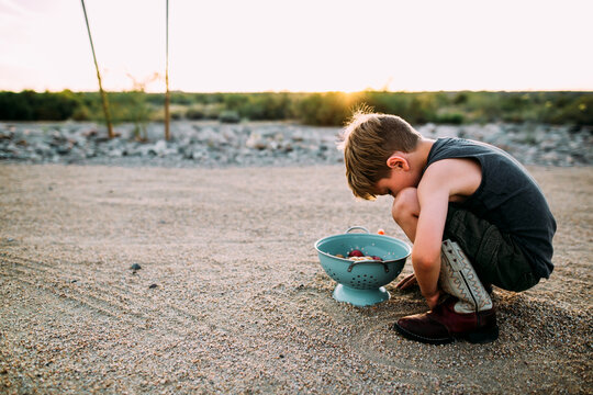 Young Boy in Cowboy Boots Kneeling over Harvest in a Blue Colander at Sunset in Desert Landscape