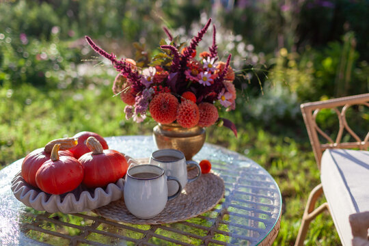 Hot coffee served in backyard on table by bouquet of fall flowers, pumpkins in morning autumnal garden. Lounge furniture