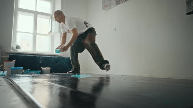 Young adult Caucasian male artist pouring blue paint from bucket onto large black canvas on floor in studio, wide shot. Clip is ideal for creative agency promos or art school advertisements