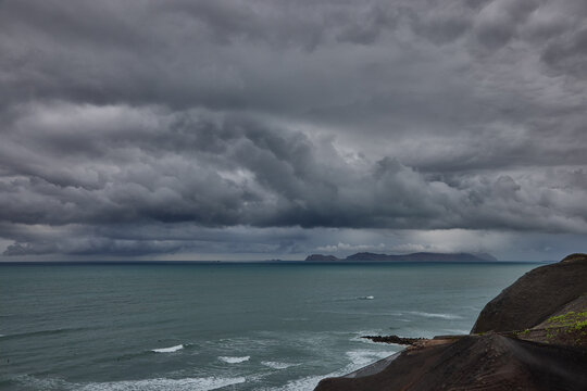 storm clouds roll over San Lorenzo Island as seen from the cliffs of Miraflores, Lima, casting a moody, atmospheric veil over the Pacific coastline. The dense cloud formations and shifting light hint 