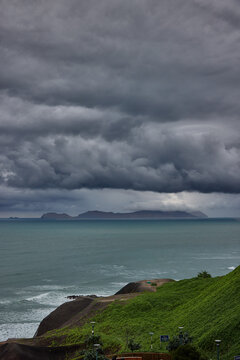 storm clouds roll over San Lorenzo Island as seen from the cliffs of Miraflores, Lima, casting a moody, atmospheric veil over the Pacific coastline. The dense cloud formations and shifting light hint 