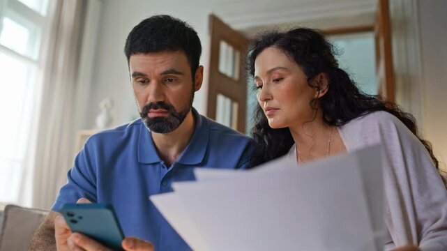 Couple checking documents phone home. Man woman reviewing bills relieved.