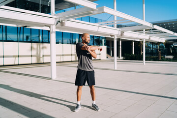 Fototapeta premium Black athlete stretching arms outdoors, muscular man warming up before workout on city plaza, healthy lifestyle, fitness motivation, authentic representation, full length, copy space, real people.