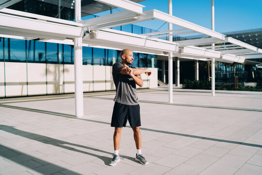 Black athlete stretching arms outdoors, muscular man warming up before workout on city plaza, healthy lifestyle, fitness motivation, authentic representation, full length, copy space, real people.