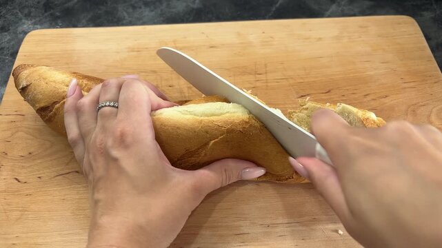 Young woman slicing a fresh baguette with a kitchen knife on a wooden board, bread preparation, home cooking, meal making.