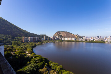 Fototapeta premium Panoramic view of Rodrigo de Freitas Lagoon in Rio de Janeiro, Brazil, with city skyline, lush green mountains, and calm waters under a clear blue sky