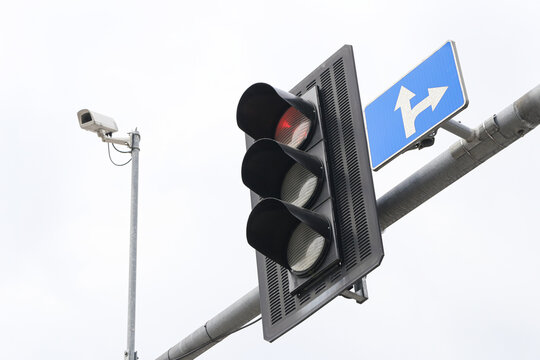 Red traffic light beside direction sign. Urban surveillance camera on metal pole. Modern road safety infrastructure. Street intersection equipment. Transportation technology background.