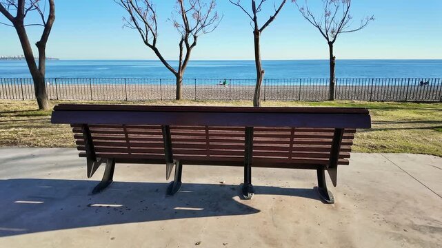 Empty wooden park bench faces the calm blue sea with bare trees and fence in antalya.