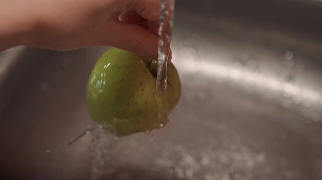 Hand holds green apple under running water washing it in sink. Stream of water flows. Stainless steel kitchen faucet with metallic drainboard with apple washing water sink