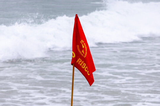 Red danger warning flag on a sandy beach with rough sea waves and white foam in the background