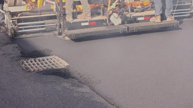 Construction workers operate machine equipment to lay new asphalt on city street at nearby drain.