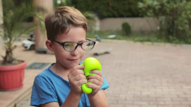 Smiling little boy wearing glasses playing with two colorful dumpling shaped squishy toys outdoors, holding sensory stress relief toys during fun playtime in the garden