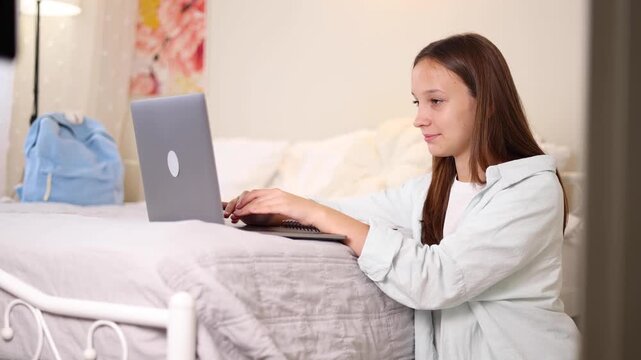 Teenage girl using laptop while lying on bed at home. Modern lifestyle, online communication and remote learning. 