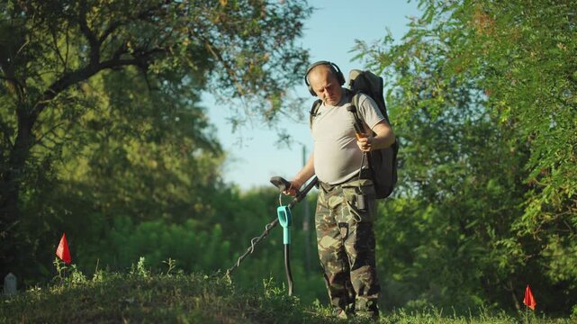 Treasure hunter with backpack uses metal detector on grassy hill surrounded by small red flags, focusing on detecting buried objects in a lush green outdoor environment under clear blue sky