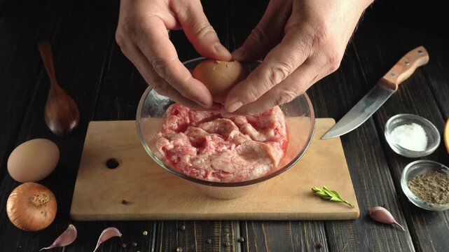 Hands of a male chef cracking eggs into a glass bowl containing minced meat, with wooden utensils, spices, and fresh ingredients arranged on a dark wooden countertop in a kitchen setting
