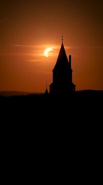 Vertical drone footage of Bouzov Castle in silhouette with a partial solar eclipse setting behind the tower in warm evening light, Czech Republic. Colors: orange, black, yellow.
