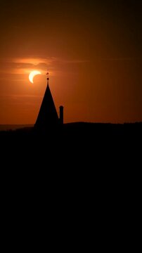 Vertical drone footage of Bouzov Castle in silhouette with a partial solar eclipse setting behind the tower in warm evening light, Czech Republic. Colors: orange, black, yellow.