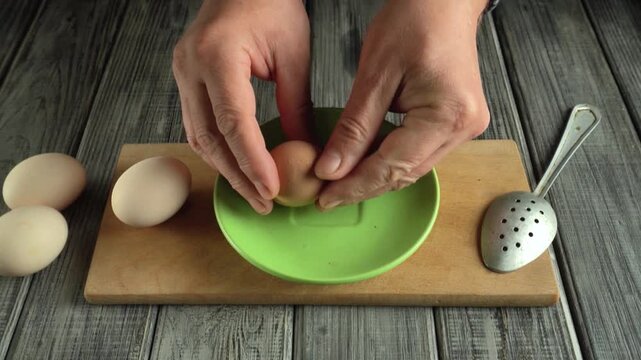 Hands cracking brown egg into green plate using a spoon, with additional eggs on wooden cutting board and rustic wooden background enhancing the kitchen setting