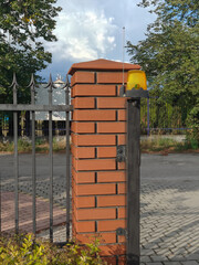Fototapeta premium Automatyczna brama wjazdowa z sygnalizatorem świetlnym i anteną sterującą zamontowanymi na ceglanym słupku ogrodzeniowym.