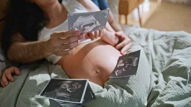 Closeup couple holding sonogram photos over pregnant belly. Ultrasound on bed.